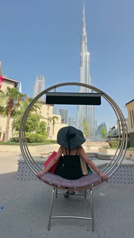 Stylish shopping woman relaxes sitting on a stylish circular chair with shopping bags, enjoying the ambiance of iconic backdrop of the Burj Khalifa skyscraper in Dubai downtown, no logos