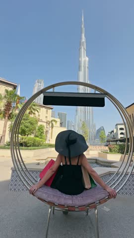 Stylish shopping woman relaxes sitting on a stylish circular chair with shopping bags, enjoying the ambiance of iconic backdrop of the Burj Khalifa skyscraper in Dubai downtown, no logos