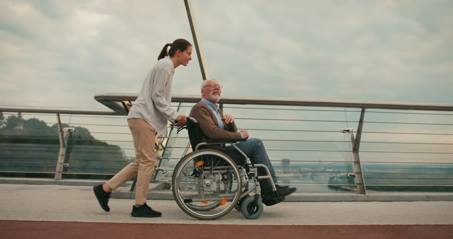 Woman walks with elderly man in wheelchair on bridge against cityscape talking. Man points out with interest to companion suggesting moment of sharing