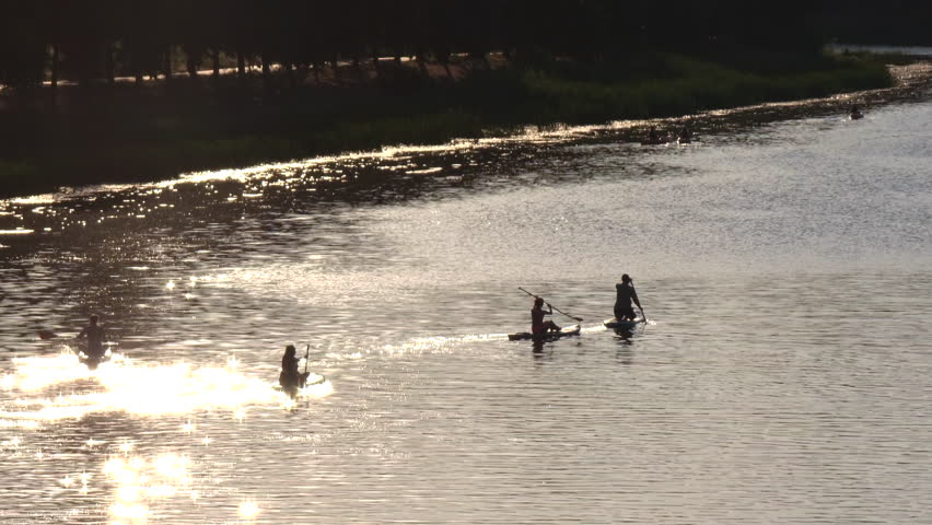Golden sunlight glimmering across river surface, silhouetted athletes paddling during serene evening training session, capturing dynamic watersport moment with scenic landscape backdrop