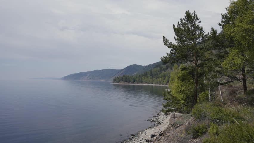 Rocky shore of lake Baikal bordering dense forest, distant mountain silhouettes rising against cloudy sky, capturing serene wilderness of Siberian landscape. Concept of relax, travel, freedom.