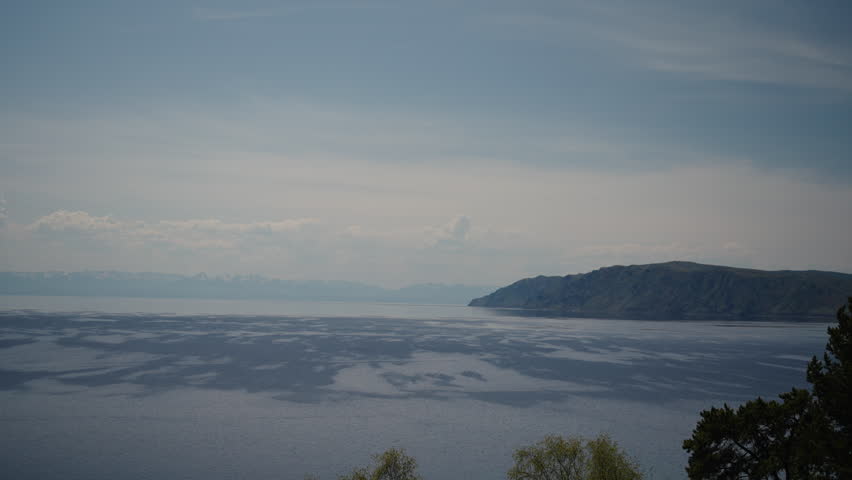Panoramic scenery view of calm lake Baikal mirroring cloudy skies, snow-dusted Sayan mountains rising beneath soft summer sunlight in pristine siberian wilderness, no people, nobody, slow motion.