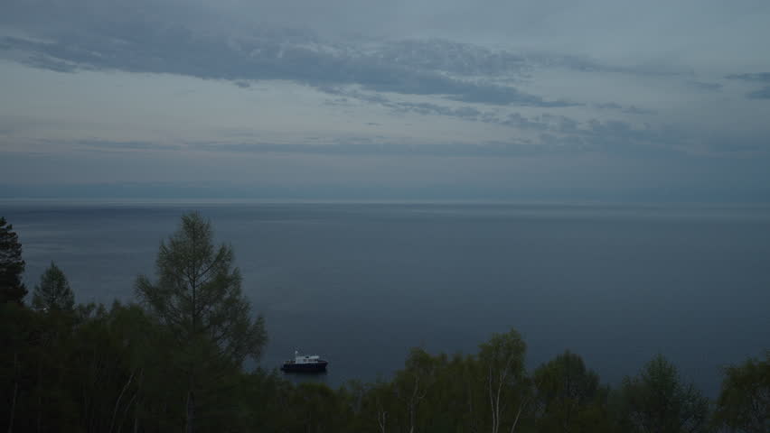 Tranquil scene of lone boat sailing on vast expanse of lake Baikal at dusk, with shoreline trees framing view and cloudy sky overhead creating serene atmosphere, no people, nobody, slow motion.