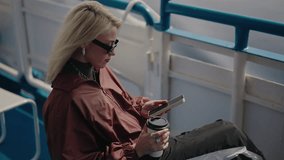 Young blond woman wearing stylish sunglasses and casual attire, sipping coffee and using smartphone while enjoying relaxing boat trip across serene lake. Shooting in slow motion. - Powered by Shutterstock - Get 15% off with code: PIKWIZARD15