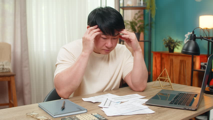 Asian man sitting at home table grips head, unpaid bills scattered with calculator and laptop, shoulders droop. Chinese guy sighs, showing financial stress and urgently seeking workable answer now.