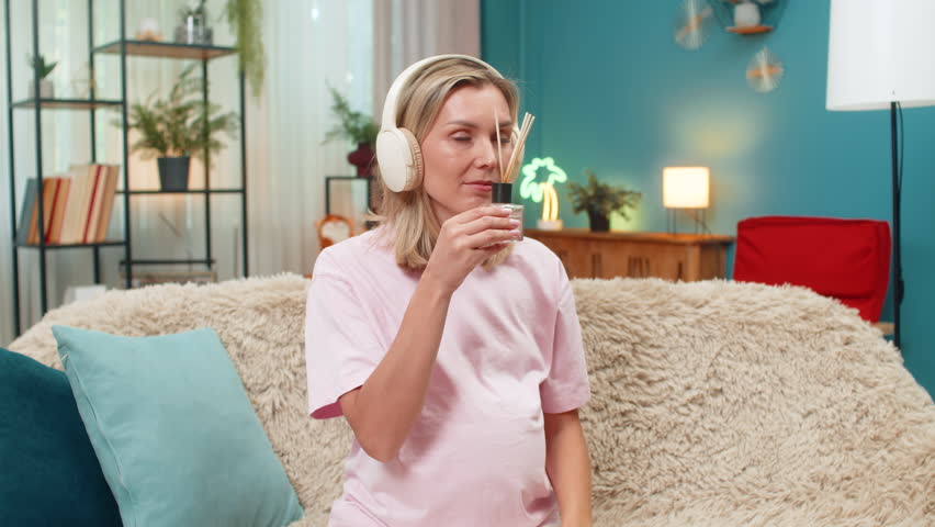 Pregnant middle-aged woman at home inhales aroma vapor from diffuser eyes closed headphones playing calm music. Caucasian girl on sofa hugs belly breathing slowly enjoying relaxing scent mood peace.