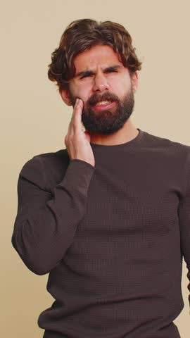 Indian bearded man touching sore cheek suffering from toothache cavities or gingivitis waiting for dentist appointment gums disease. Handsome Hindu guy indoors studio shot isolated on beige background