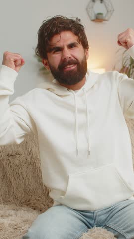 Happy Indian young man watching TV shouting, celebrating success, winning, goal achievement good news, lottery luck, victory sitting on sofa in living room. Adult guy clenching fists in home room.