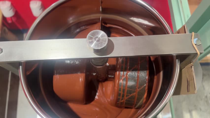  close-up video of melted chocolate being mixed in a professional chocolate melanger, with smooth, rich chocolate swirling under rotating granite rollers inside the stainless steel machine.
