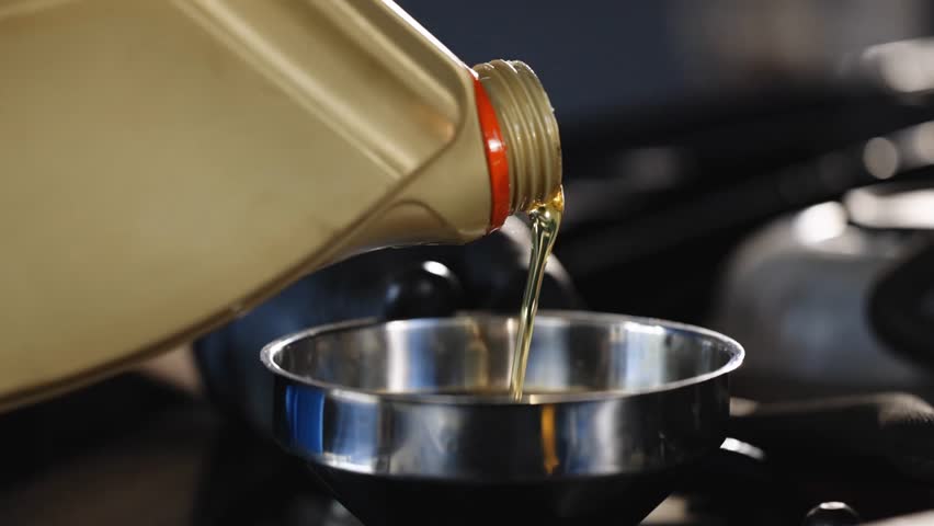A close-up shot of a mechanic pouring oil from a plastic container into the engine through a funnel, performing routine maintenance on a vehicle.
