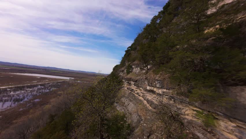 High-speed drone fly along of Southern Illinois river bluffs, skimming trees on the cliff edge under a sunny spring sky with vibrant blue tones. Perfect for looking like a bird