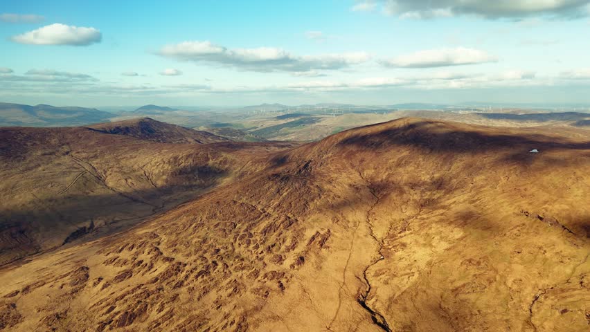 A scenic look at the mountainous terrain around Priest's Leap in County Cork, Ireland. Wind turbines are visible on the horizon, providing a distant contrast to the natural landscape.