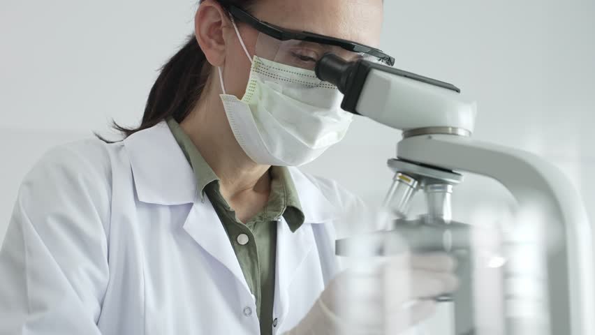 Female scientist examining microscope slide, wearing protective gear while conducting medical research in sterile laboratory environment. Medicine, healthcare and science concept - Powered by Shutterstock - Get 15% off with code: PIKWIZARD15