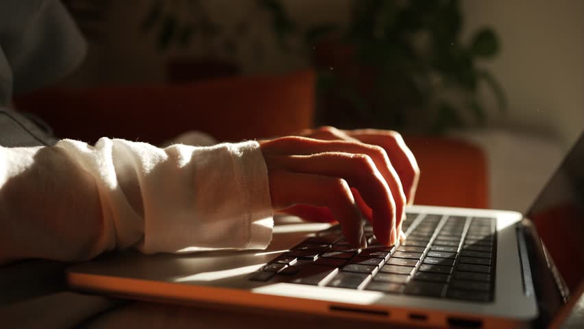 Sunlight illuminates woman's hands as she diligently types on her laptop keyboard, working remotely from the comfort of her home, creating a serene and productive atmosphere