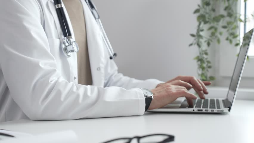Doctor man wearing a white coat and stethoscope is typing on a laptop in a bright medical office, showcasing the intersection of healthcare and technology. Medicine and health care - Powered by Shutterstock - Get 15% off with code: PIKWIZARD15