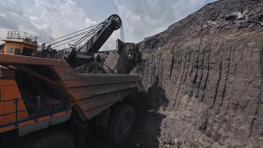 FPV drone view work on mining industry from above. Big excavator removes overburden soil and loads it into large yellow dump truck at coal quarry.