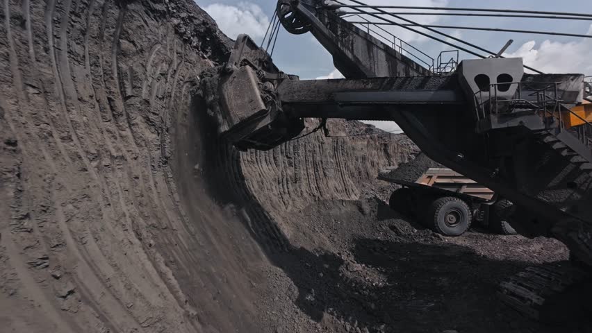 Loading process overburden soil from big excavator into large yellow dump truck at coal mining quarry, FPV drone view.