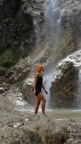 Beautiful woman walking around Alps waterfall with water going up. Fun reverse slow motion shot
