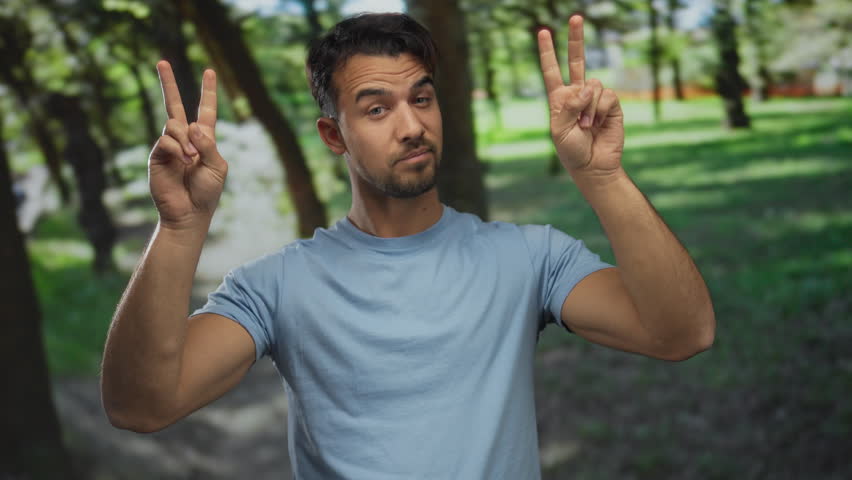 Young hispanic man in a park gesturing with finger quotes amid greenery, conveying sarcasm and humor while wearing a casual blue shirt outdoors.