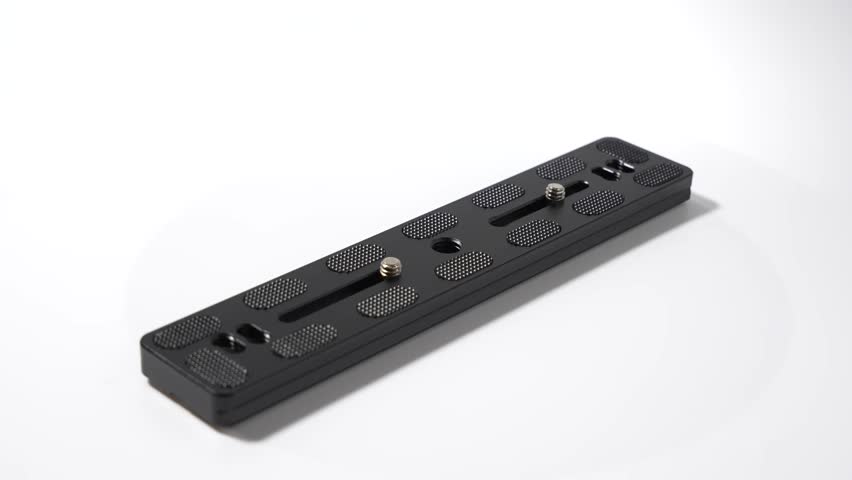 Close-up of a black metal quick-release plate with textured grips and mounting screws. The plate rests on a clean white background.