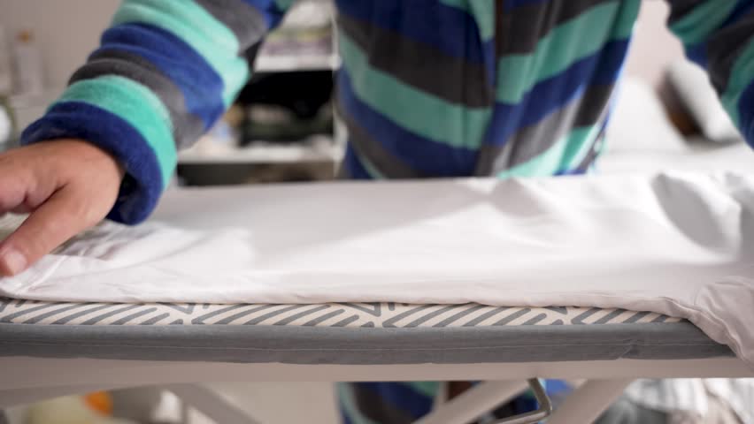 Close-up of steam iron pressing a white shirt. The patterned ironing board contrasts with the plain fabric.