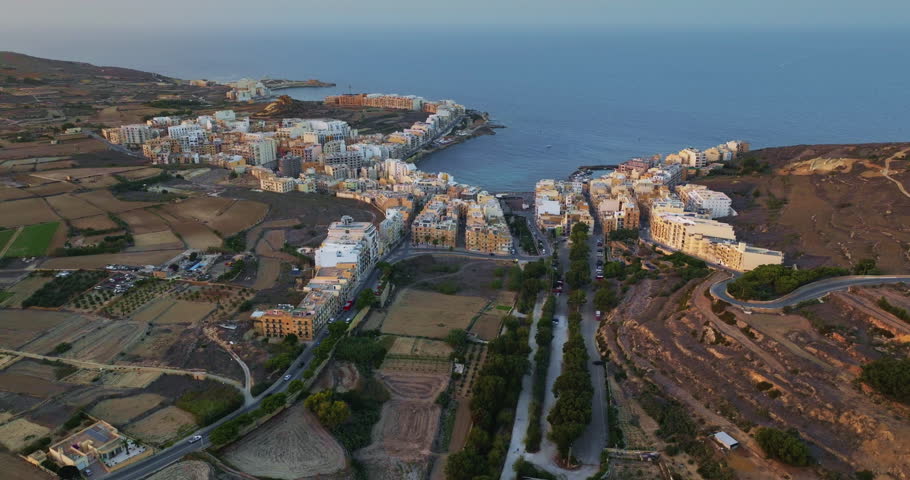 Malta, Comino island: Marsalforn resort in Gozo, revealing the urban modern city buildings landscape, agricultural fields, and the tranquil Mediterranean Sea. Drone flight footage aerial view