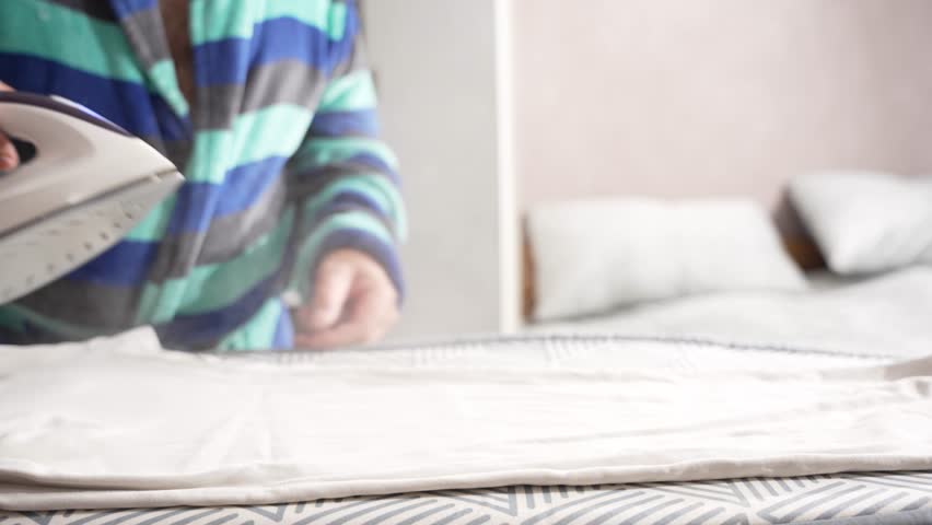 A striped-sleeve hand holds a steam iron over a white shirt. The ironing board is neatly set up in a bedroom.