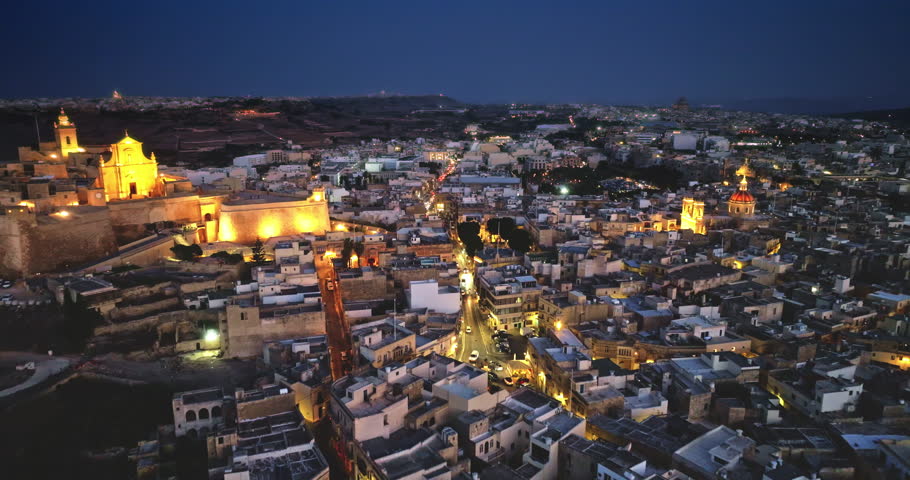Malta, Comino island: Victoria, Gozo's capital, historic charm and vibrant nightlife with illuminated road traffic, church and buildings glowing against the twilight sky. Drone flight footage