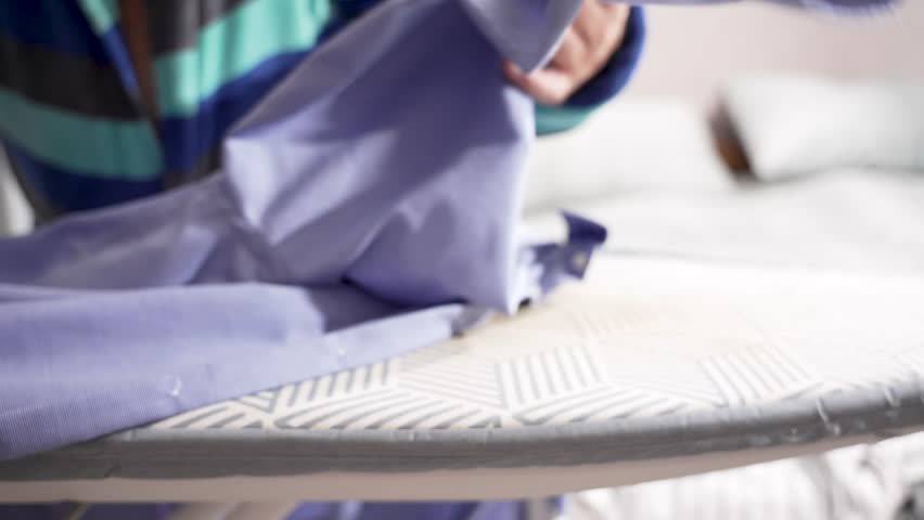 A blue shirt is ironed on a patterned board, with hands holding fabric taut. Steam gently smooths the surface.