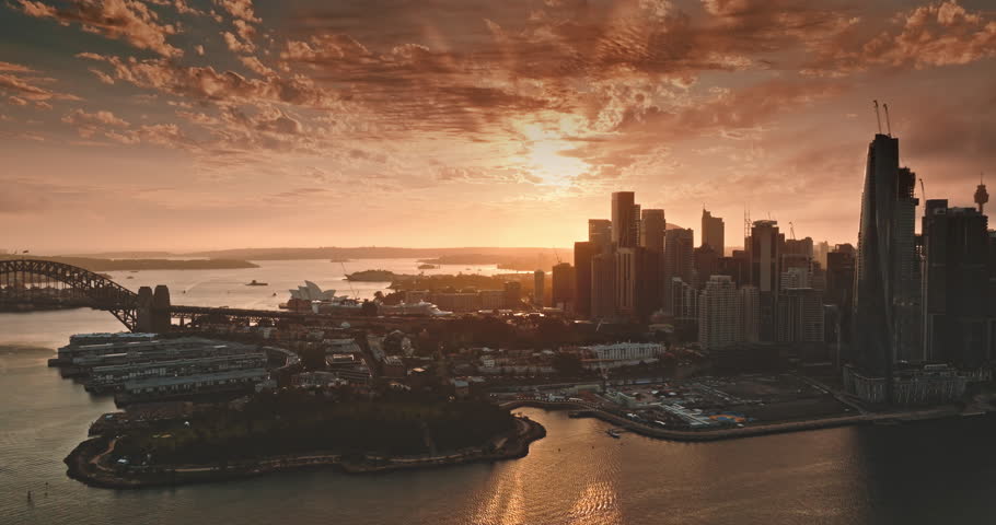 Australia, Sydney: Warm sunset over Sydney skyline, reflecting on water and showcasing Sydney Harbour Bridge and Opera House, creating a beautiful cityscape landscape. Drone flight footage aerial view
