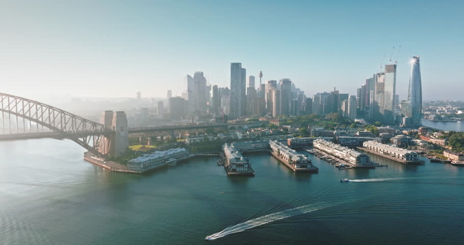 Australia, Sydney: Breathtaking aerial view of Sydney Harbour at sunrise, highlighting the Sydney Harbour Bridge, modern city skyline, and bustling boats navigating the water. Drone flight footage
