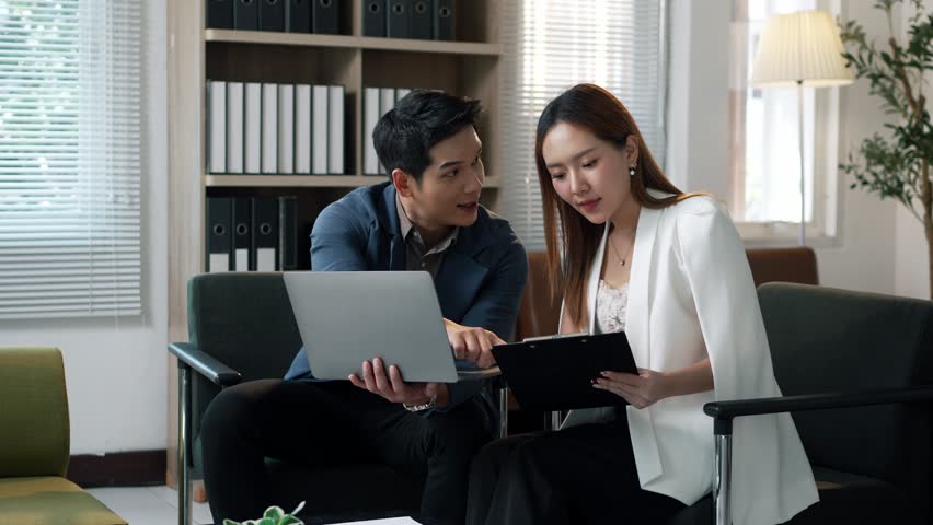 Corporate professionals collaboratively reviewing financial data on laptop, discussing project strategy while sitting together on office sofa with engaged, confident demeanor