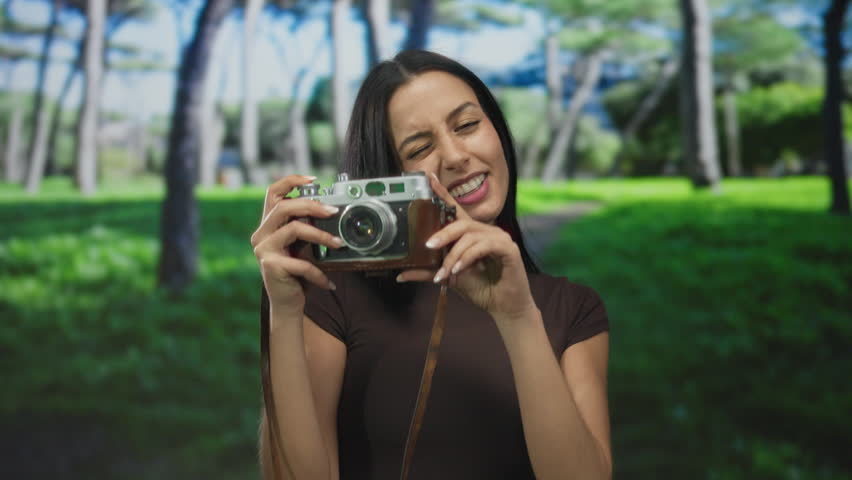 Woman smiling with vintage camera in park under sunlit trees showcasing blissful outdoor photography moment.