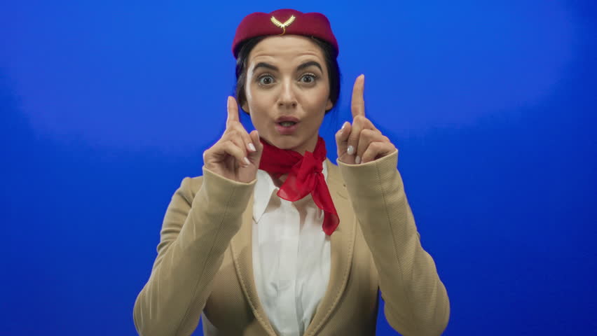 Young woman flight attendant in uniform makes inviting gesture against blue wall background expressing friendly hospitality smiles brightly