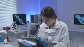 Young female scientist in lab coat taking notes while using microscope, with laptop and test tubes nearby in modern laboratory with DNA double helix graphic displayed on screen - Powered by Shutterstock - Get 15% off with code: PIKWIZARD15