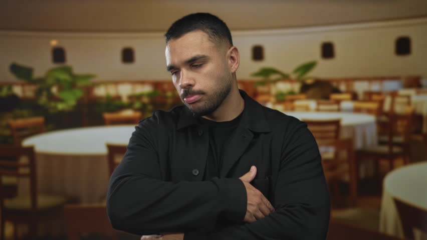 Man in black jacket crossing arms in a restaurant building amid wooden tables and chairs; quiet reflection.