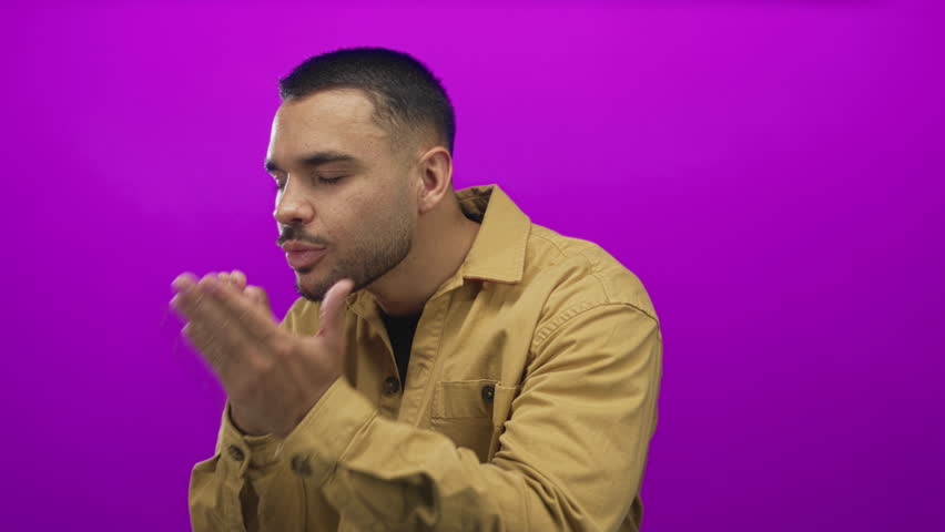 Young hispanic man in brown shirt closes eyes and blows kiss against magenta studio backdrop; affection warmth.
