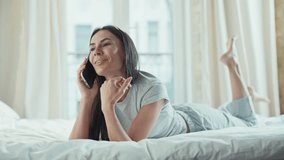 Happy Caucasian woman lying on bed holding smartphone to ear with closed eyes smiling. Talking cheerfully during phone call. Enjoying pleasant conversation while relaxing in bright bedroom. - Powered by Shutterstock - Get 15% off with code: PIKWIZARD15