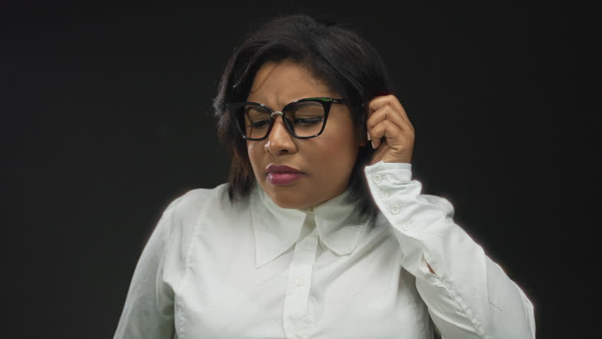 Woman wearing glasses on black background gesturing to listen intently with hand to ear, expression showing curiosity and focus.