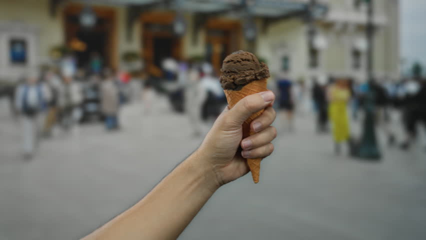 Man holding chocolate ice cream cone on a bustling city street as people walk by in the background, highlighting urban outdoor indulgence.