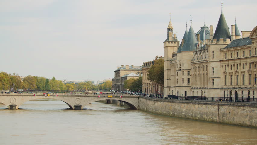 Paris, France -- October 10, 2024. Historic view of La Conciergerie along the Seine River in Paris, with its gothic towers, arched stone bridge, and tree-lined embankment on a calm autumn day.