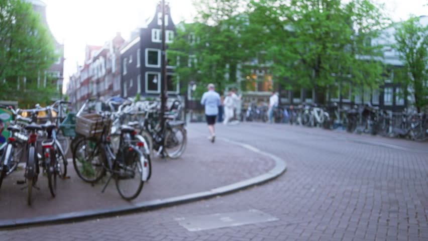 Bicycles lining a curved street in amsterdam, netherlands, with blurred people walking and cycling among traditional canal houses in a soft focus urban scene