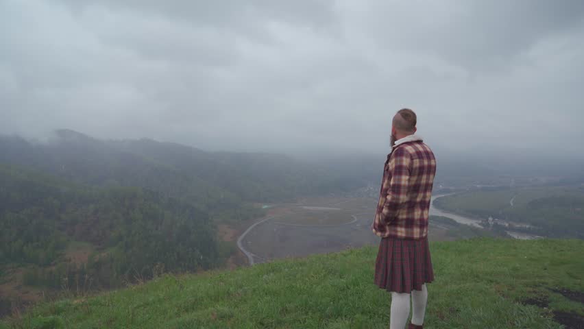 An adult redhead bearded man in Scottish national dress plaid jacket and kilt stands on a hill and looks into the distance. Masculinity, pride and human dignity concept.