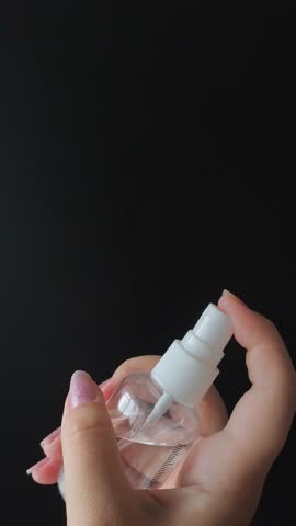 A woman's hand presses the button of a transparent plastic spray bottle and sprays water on a black background. Spray, perfume.