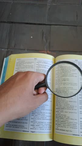 PAMEKASAN, INDONESIA - AUGUST 16, 2025: An Asian hand holds a magnifying glass, searching for the words "wedding" and "war" in an English-Indonesian dictionary. A concept exploring contrasting themes.