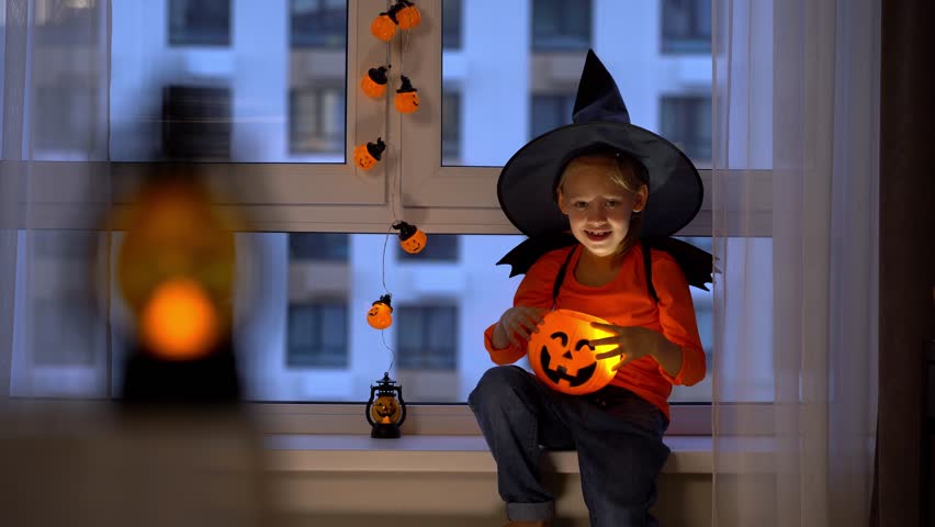 Happy girl sits by window in witch hat and conjures over halloween pumpkin lantern in evening. Child celebrates halloween