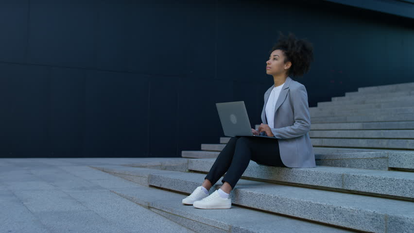 Young professional woman sits on outdoor steps with a laptop, looking away thoughtfully while considering an important decision about her future.