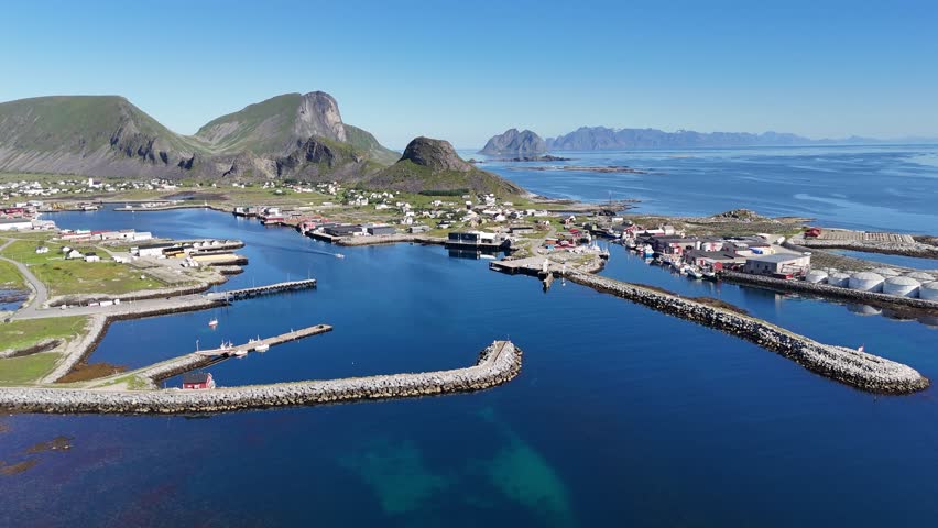 Aerial view of Værøy harbor: colorful village by the sea, dramatic mountains rising behind, and the endless blue of the Norwegian coast.