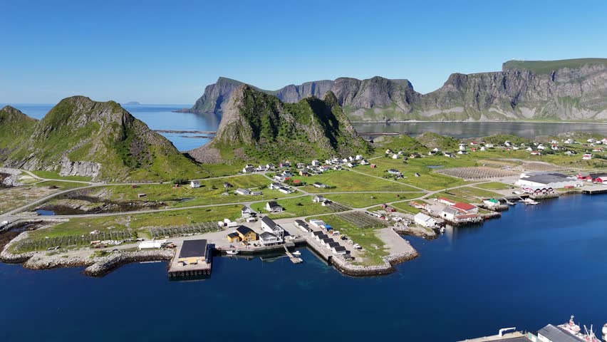 Aerial view of Værøy harbor: colorful village by the sea, dramatic mountains rising behind, and the endless blue of the Norwegian coast.
