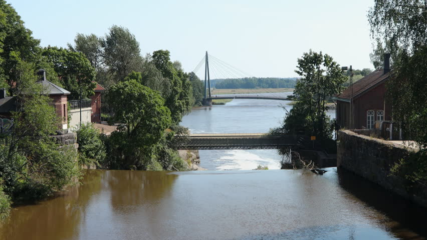 View from Vanhankaupungin Lukkosilta bridge in Helsinki, overlooking the river, rapids, and historic red-brick buildings surrounded by summer greenery.
